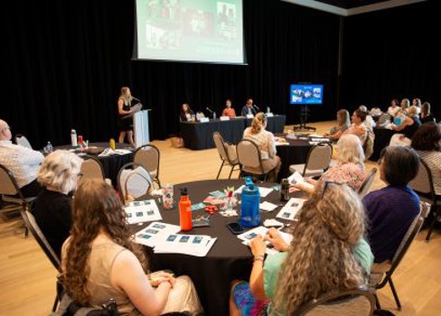 A group of people sitting at a table with a person standing in front of a screen.